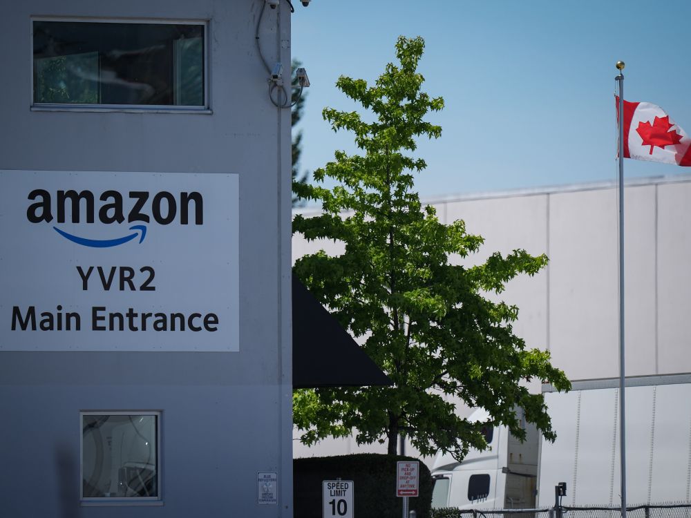 An exterior photo of a large warehouse with a sign identifying it as &ldquo;Amazon YVR2 Main Entrance.&rdquo; A Canadian flag flies nearby.