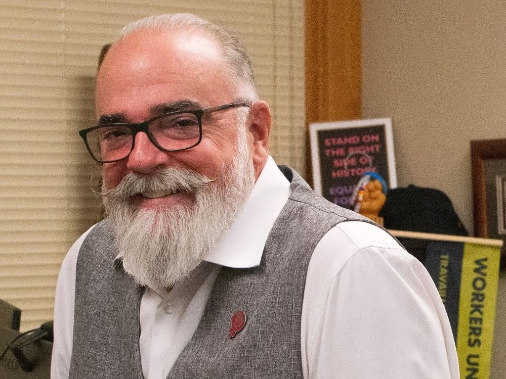 A man in his 60s with light skin and a grey moustache and beard. He wears glasses and smiles at the camera.