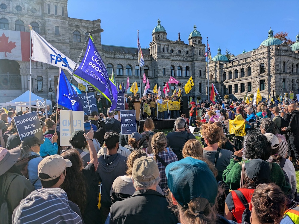 A crowd of people holding flags and banners stand in front of a stage set up outside the B.C. Legislature.