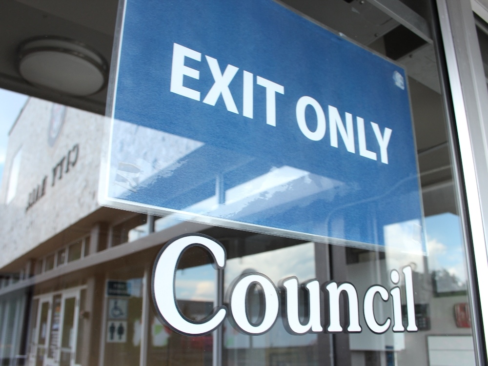 A blue laminated sign on a glass door says &lsquo;Exit only&rsquo; in white sans-serif typeface above the word &lsquo;Council&rsquo; in white text on the door. A reflection of a grey building labelled &lsquo;City Hall&rsquo; is visible on the glass.