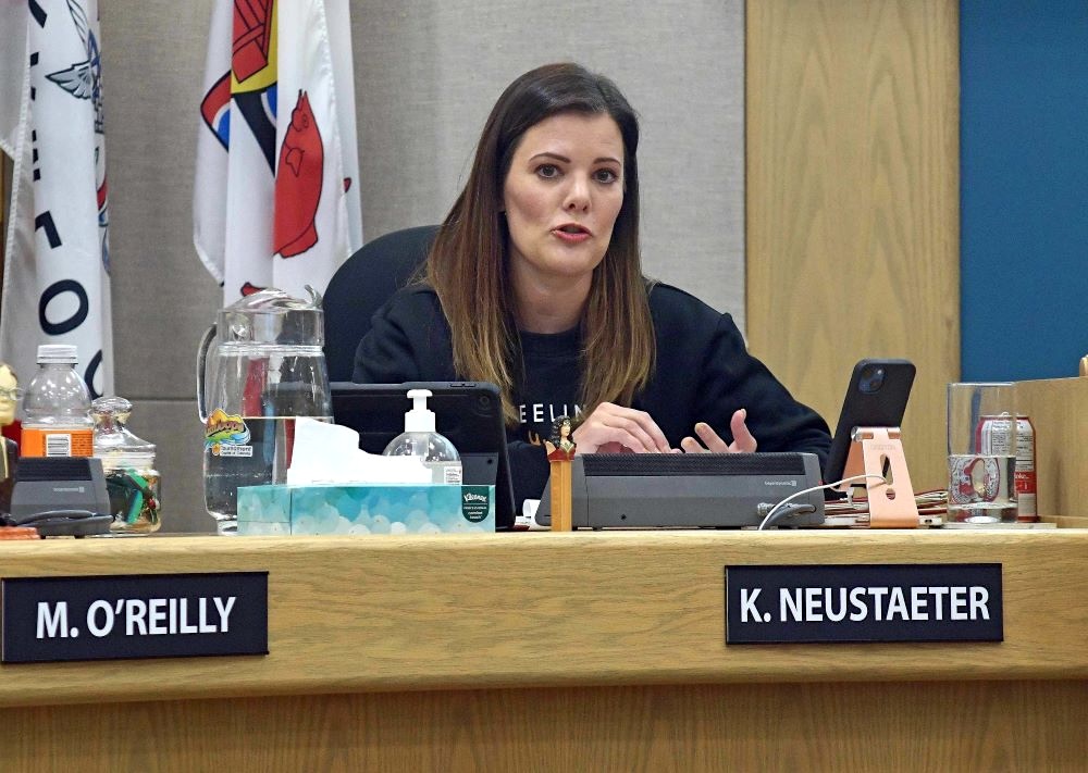A woman with light skin tone and long brown hair sits at a council chamber table behind a sign that reads, &lsquo;K. Neustaeter.&rsquo;