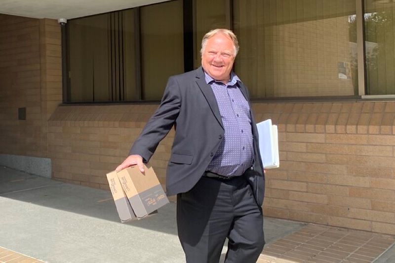 A middle-aged man with a florid light skin tone wearing a dark suit walks outside a building holding boxes in one hand and a binder in the other.