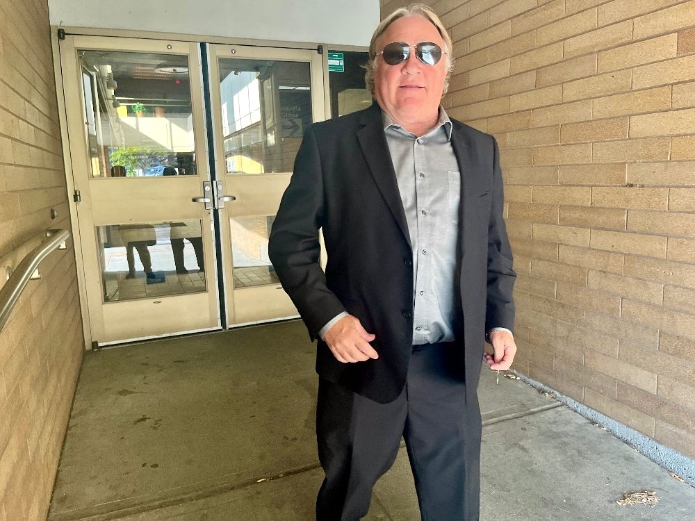 A middle-aged man with a florid light skin tone and wearing a black suit exits a courthouse.