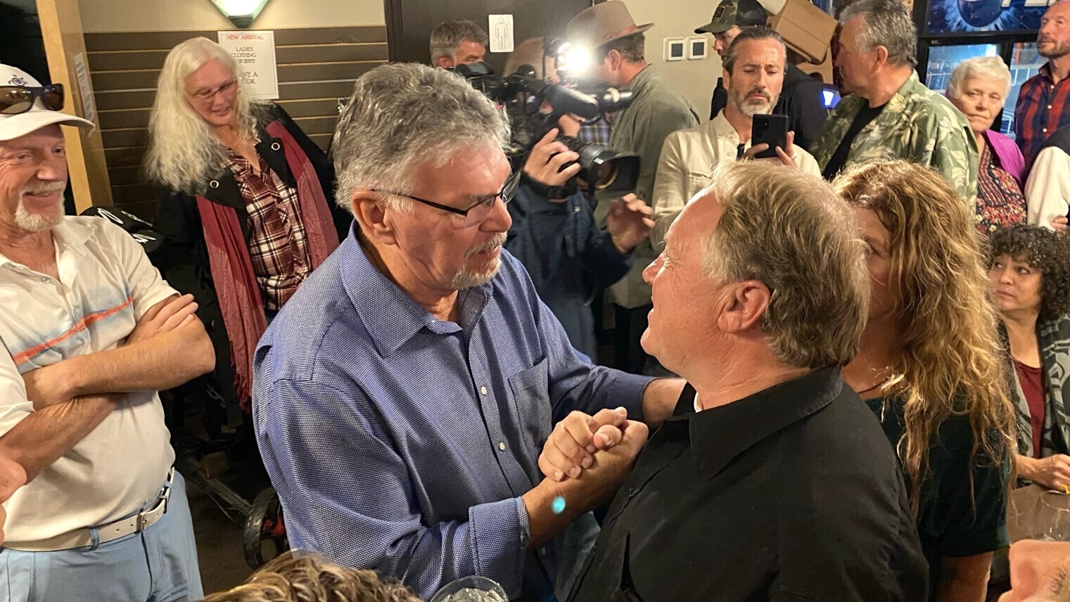 Two light-skinned middle-aged men clasp hands amid a press of people.