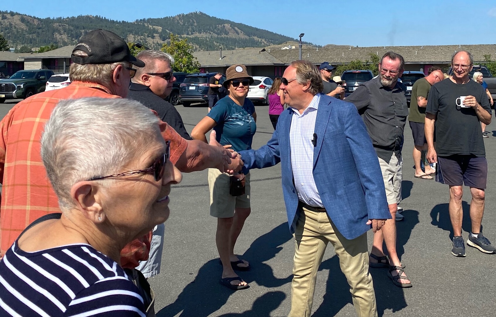 Reid Hamer-Jackson, wearing a blue jacket, beige pants and sunglasses, shakes hands with a group of smiling people outside.