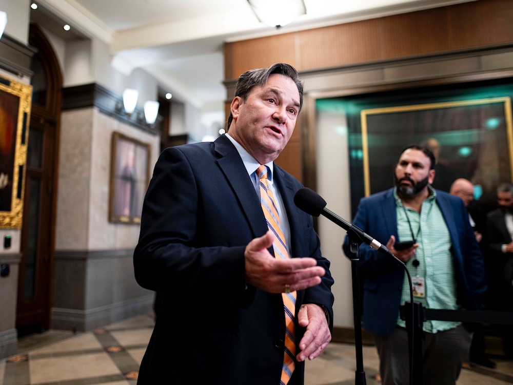 A middle-aged man wearing a suit with a striped orange and grey tie speaks to reporters in a hallway inside Canada&rsquo;s Parliament building.