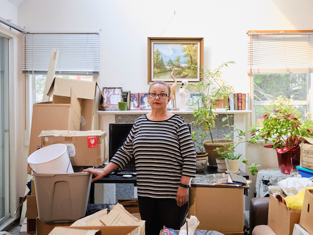 A woman in her 70s stands in the living room of her apartment, surrounded by moving boxes. She wears glasses, black pants and a black and white striped sweater.
