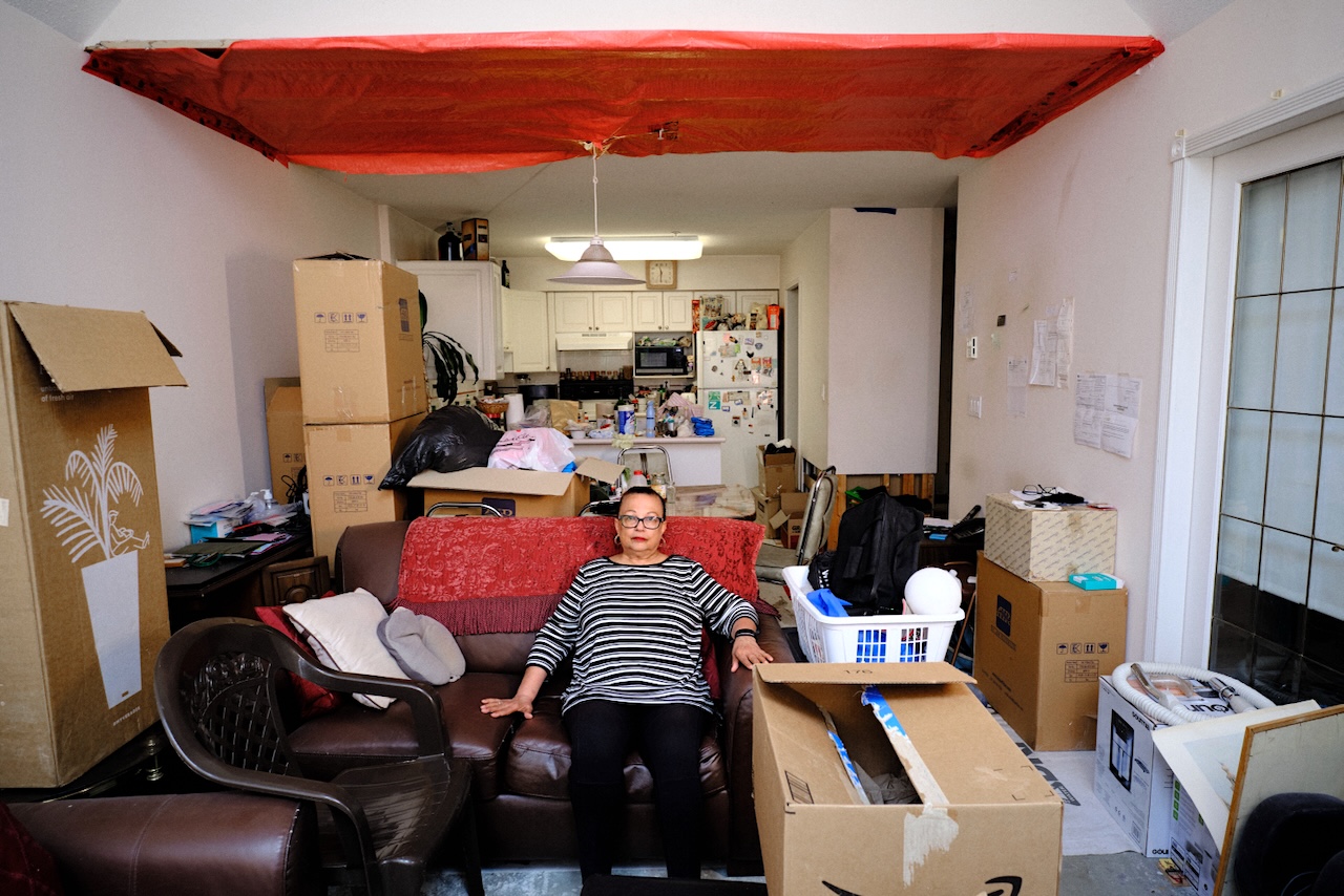 A woman in her 70s, wearing glasses and red lipstick, sits on a red couch in her living room. Above her head, a large portion of the ceiling is covered with an orange tarp.