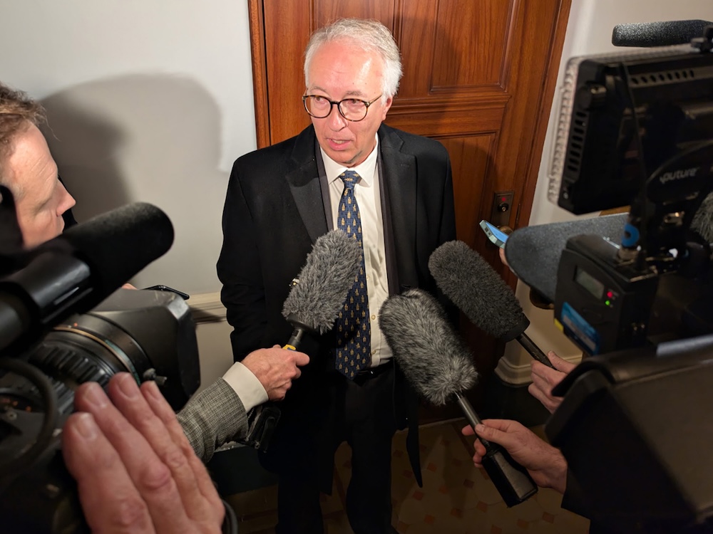 John Rustad wears a dark suit. He is ringed by members of the press holding microphones and cameras.
