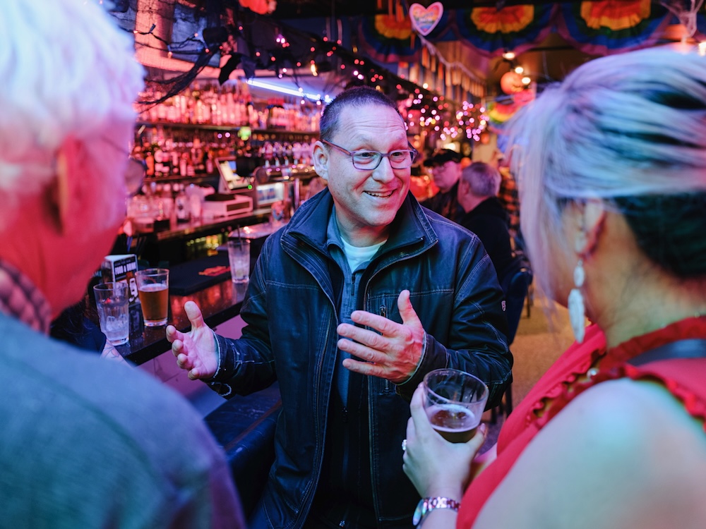 A middle-aged man with light skin wearing glasses speaks in an animated way to two other people in a bar.