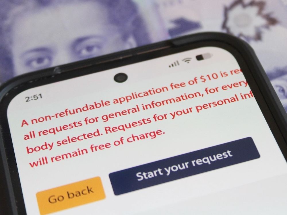 A smartphone screen shows the message &lsquo;A non-refundable application fee of $10 is required for all requests for general information for every body selected. Requests for your personal information will remain free of charge.&rsquo;