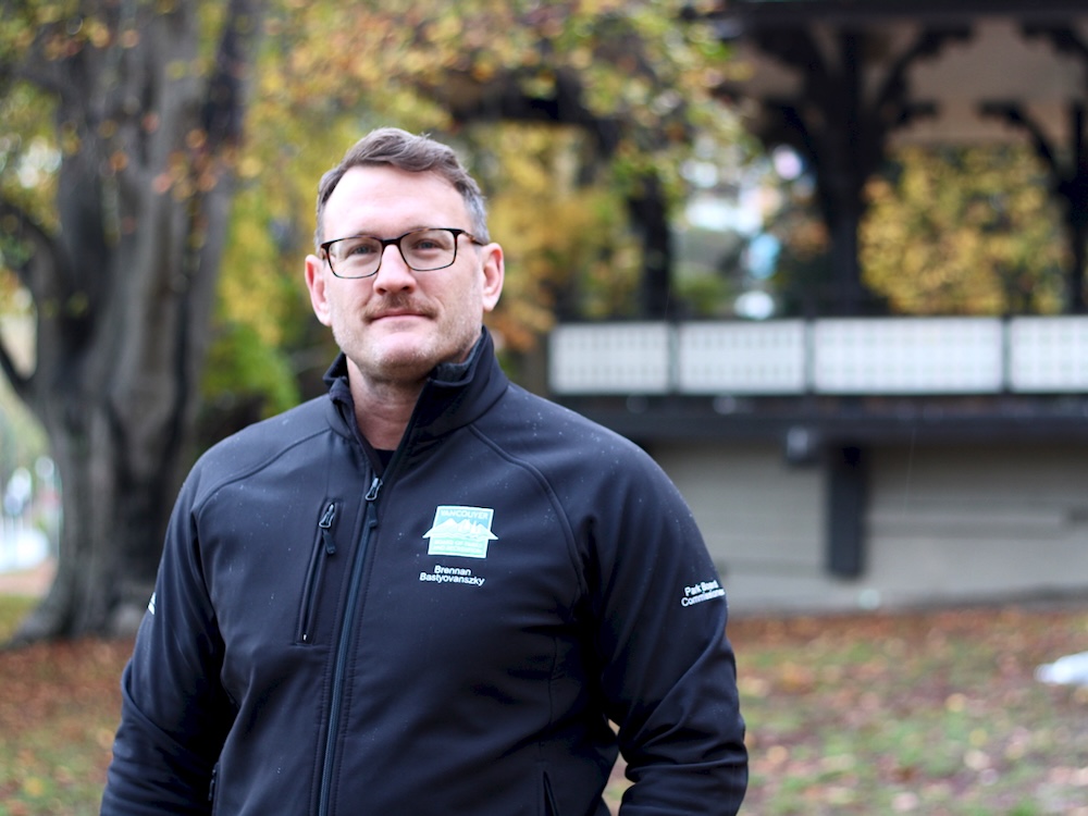 A man with light skin, brown-grey hair and a trim, brown moustache, wearing glasses, looks directly at the camera, standing in a fall park setting.