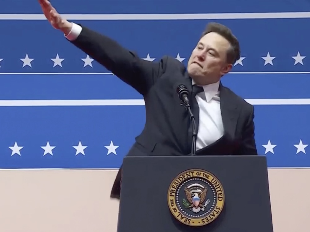 A light-skinned man wearing a dark suit stands at a podium, his right arm extended in what looks like a Nazi salute.