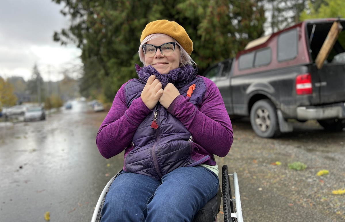 A woman with light skin tone and light-pink-coloured hair is seated in a wheelchair. She&rsquo;s wearing a yellow hat, purple shirt and purple vest.