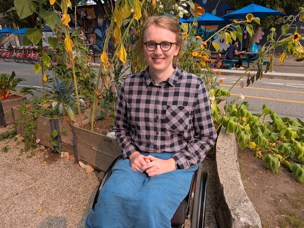 A woman with light skin and pulled-back blond hair grins at the camera while posing in front of a late-summer garden. She&rsquo;s wearing black glasses, a purple and grey flannel shirt and a blue skirt. She&rsquo;s sitting in a manual wheelchair with her hands resting in her lap.