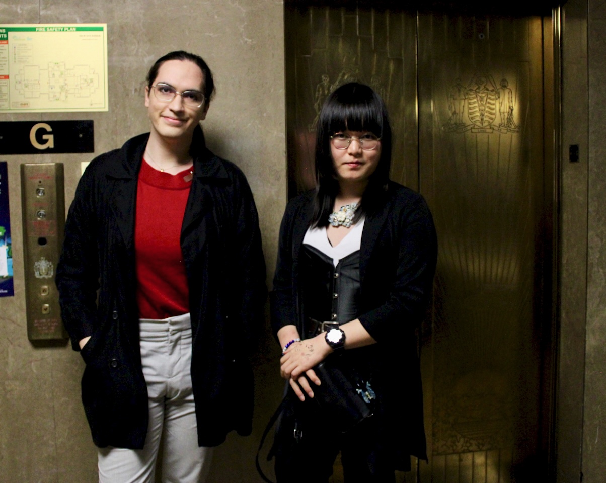 Two people, both with medium-light skin tone and long dark hair, stand by an elevator. 