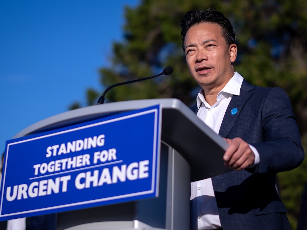 Mayor Ken Sim, a man with medium skin tone and short dark hair, speaks at a lectern that reads, &lsquo;Standing together for urgent change.&rsquo;