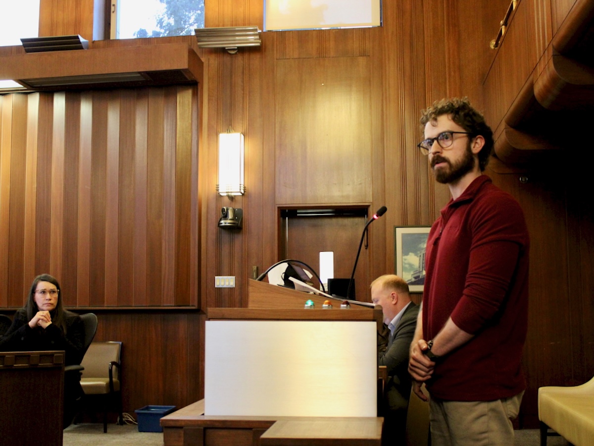 A man in his 30s with curly brown hair and glasses addresses Vancouver city council in a wood-panelled room. A seated woman is watching him.