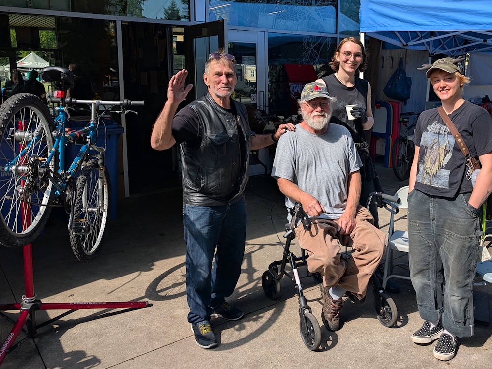 Four people wearing dark, neutral-coloured clothes stand looking at the camera with friendly expressions. A blue bike rests on a stand beside them, and they&rsquo;re standing in front of a blue tent. The man in the middle rests on a walker he helped repair.
