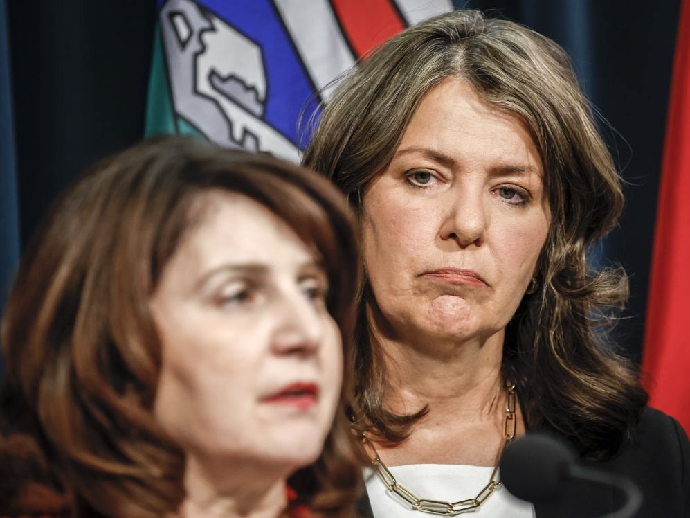Danielle Smith is a woman with light skin and wavy brown hair, and Adriana LaGrange is a woman with light skin and reddish-brown wavy hair, out of focus. Smith is watching LaGrange speak from behind, wearing a tight-lipped expression.