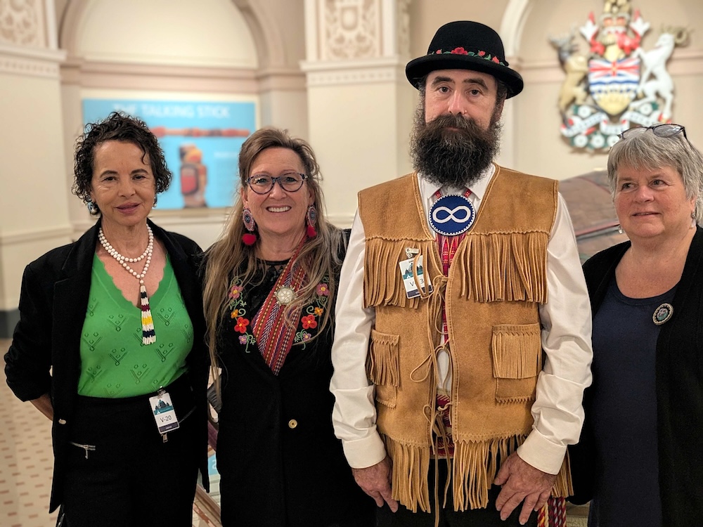 Four people, three women and a man, stand together in an ornate hall. He wears a hat and a brown leather vest with fringes.