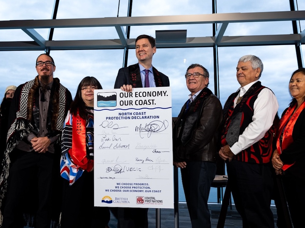 David Eby, a tall light-skinned man in suit and tie, holds a novelty cheque-sized &lsquo;North Coast Protection Declaration&rsquo; with several signatures visible on it. He is flanked by several people wearing Indigenous-patterned attire.