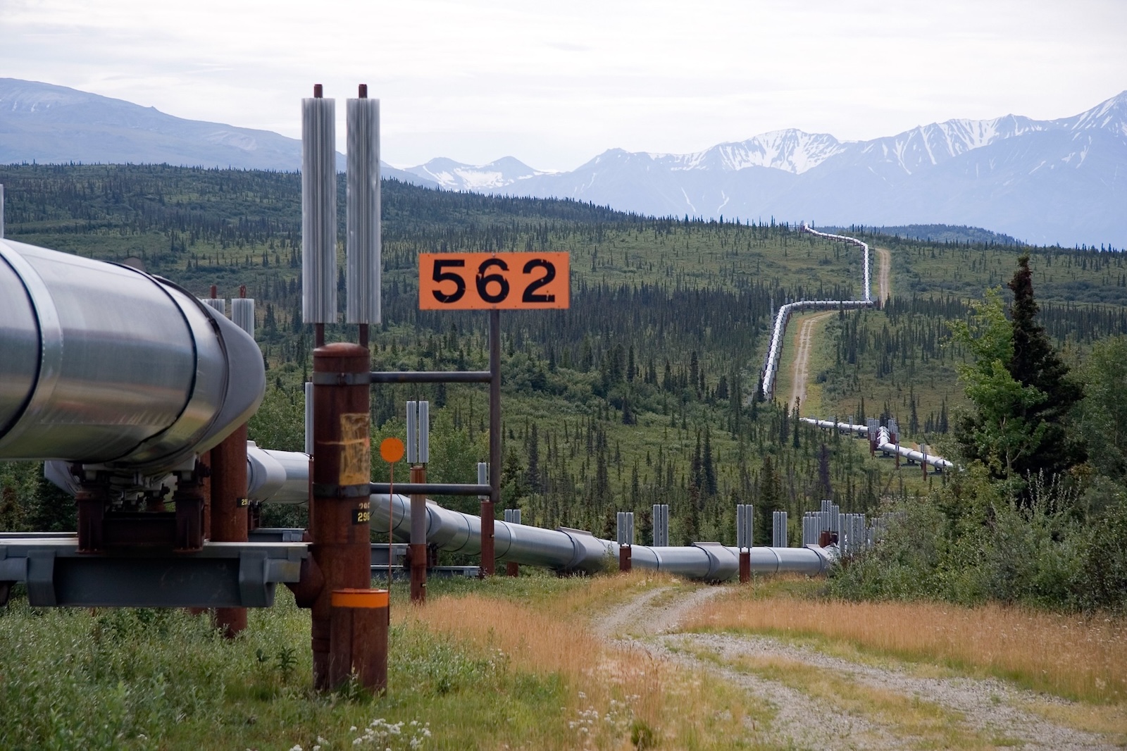 A pipeline weaves through rugged terrain with snow-capped mountains in the background.