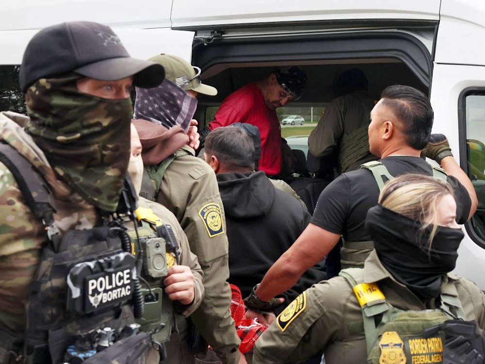 Officers in uniform, wearing masks, load two men into a van.