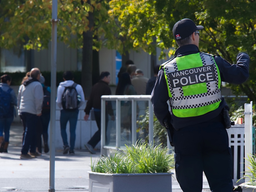 A police officer wearing a bright yellow vest with &lsquo;Vancouver Police&rsquo; across the back stands near a road. In the background several pedestrians walk away from the camera.