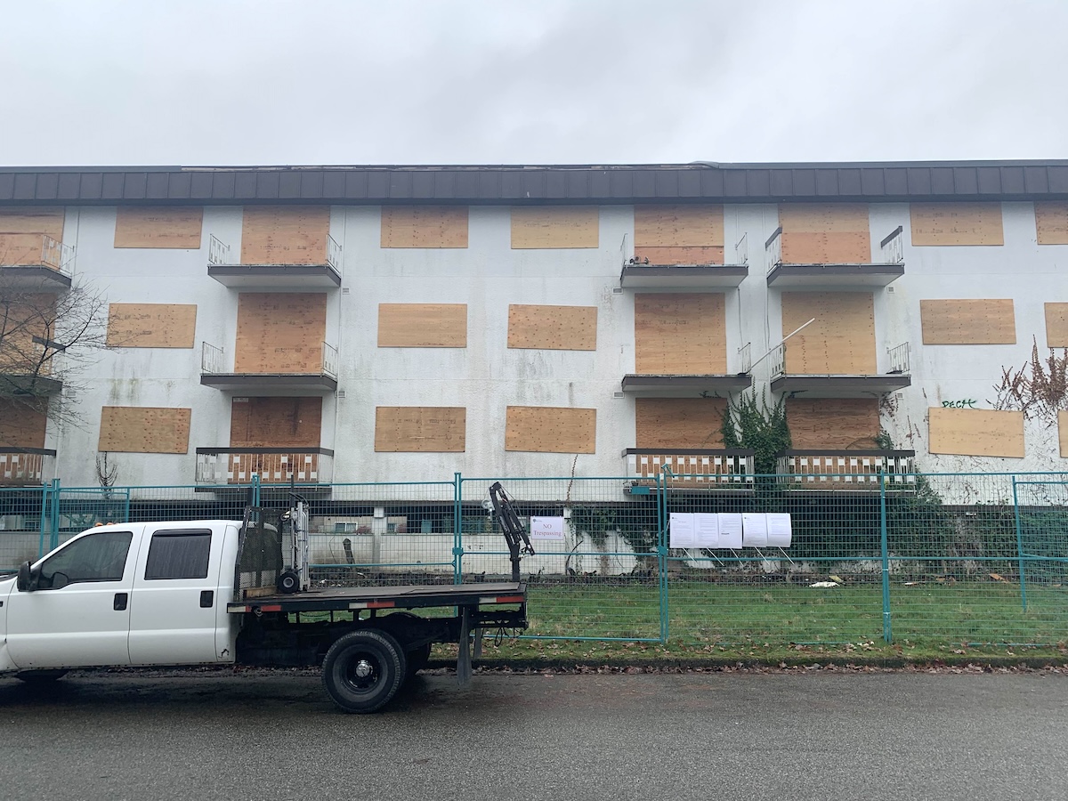 A white pickup truck drives by a wide white apartment building with its windows boarded up on a grey day.