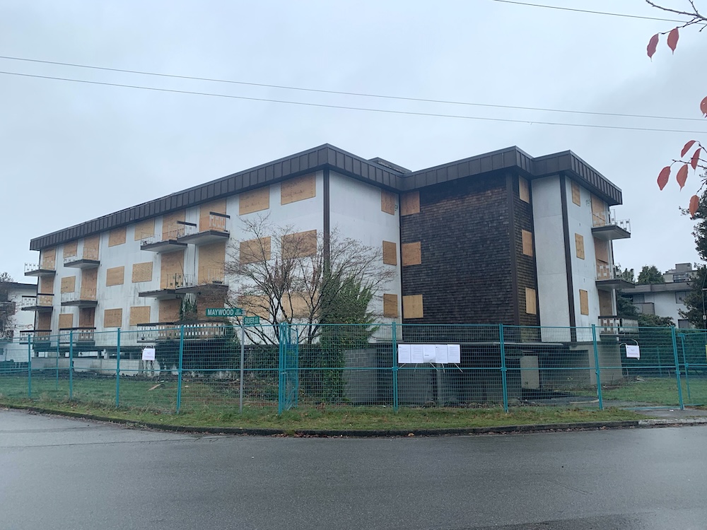 A wide three-storey apartment building with white stucco and brown brick walls has its windows boarded up with plywood. It sits on a street corner with green signs against a grey sky.