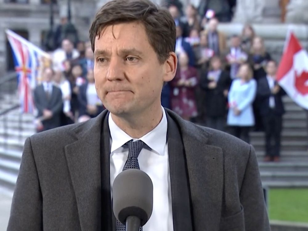David Eby stands at a microphone in a dark suit with supporters in the background.