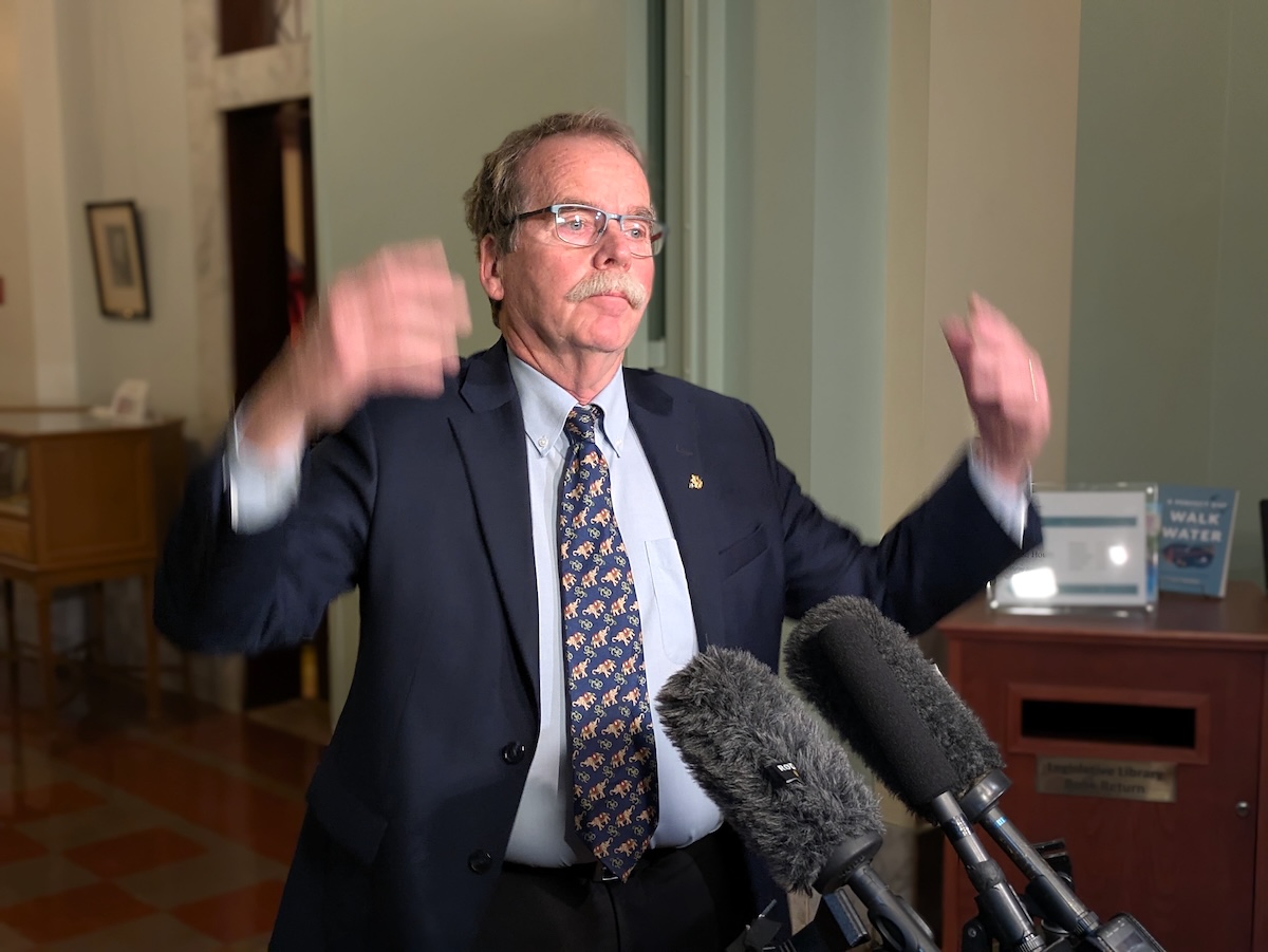 Rob Botterell, a 60-ish man in a suit, gestures as he speaks to reporters.
