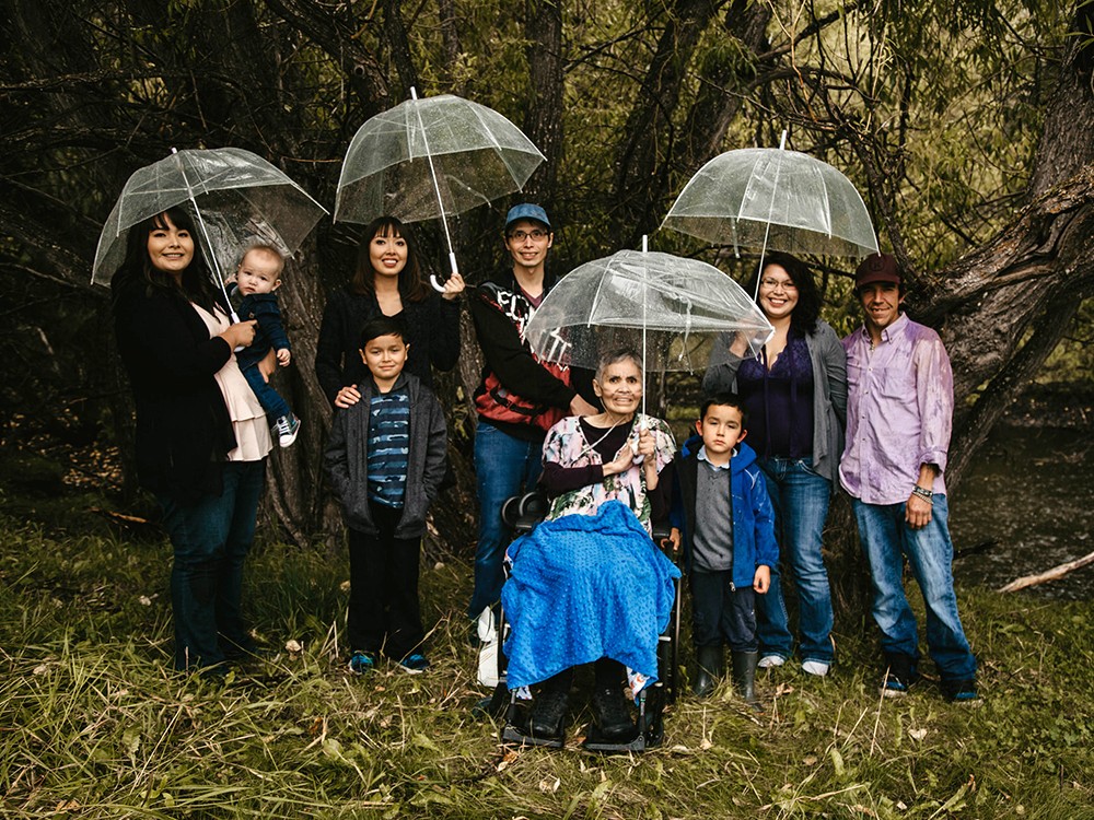 An older woman with short, greying hair and with an oxygen tube sits in a wheelchair surrounded by younger adults and children. They are in an outdoor setting and several people hold umbrellas.