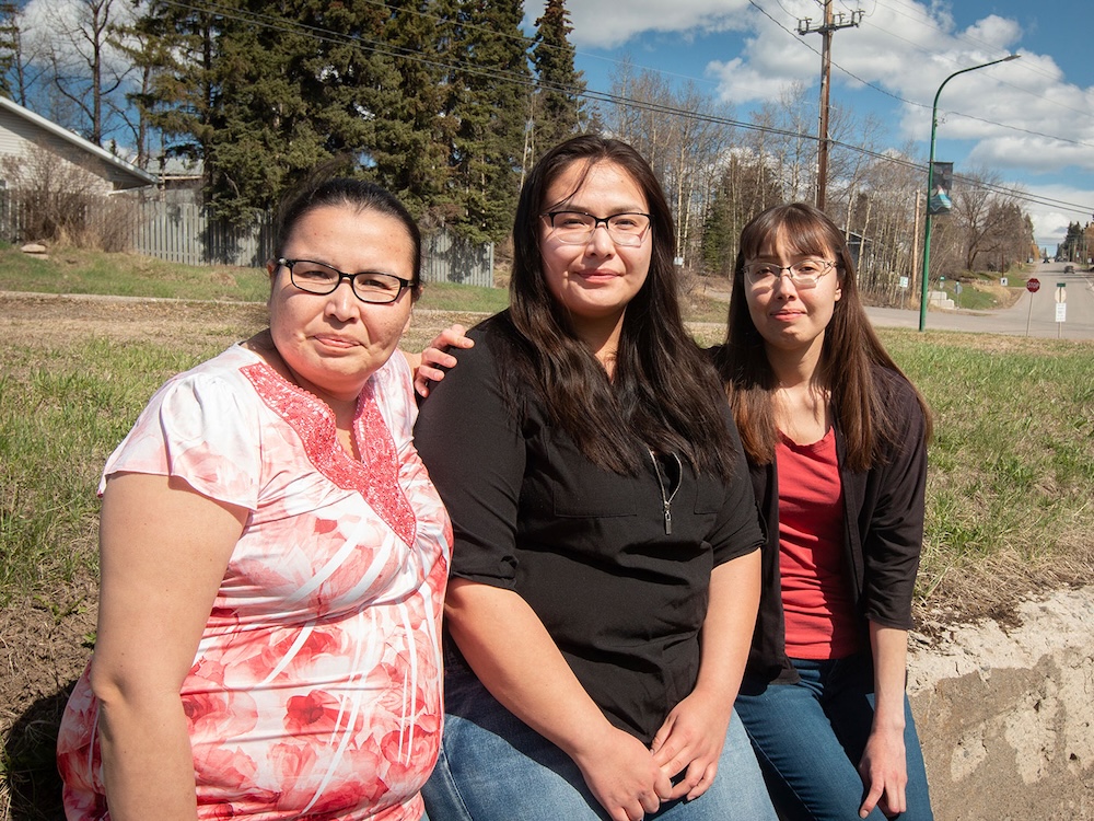 Three women, all with dark hair and wearing glasses, lean against a low wall with their arms around each other under a blue sky.