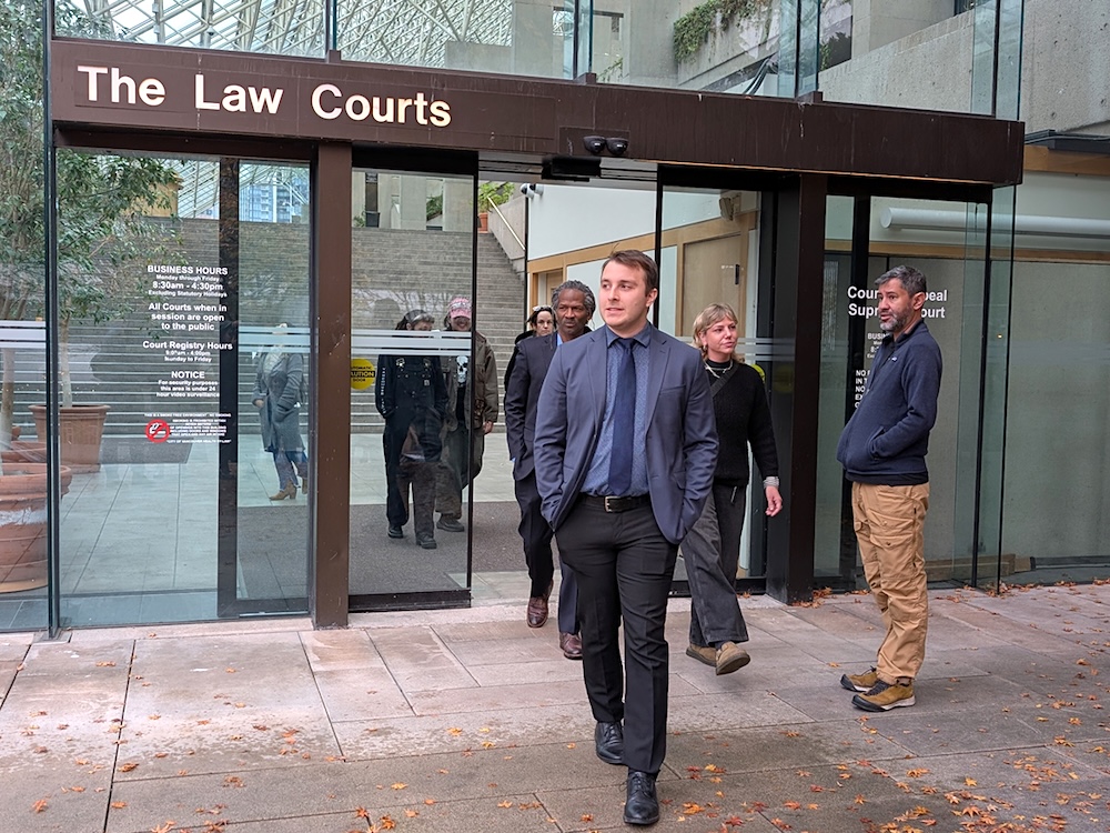 A man with light skin tone and short brown hair exits the Vancouver law courts. He is wearing black trousers, a blue collared shirt, a blue tie and a blue blazer. His hands are in his pockets.