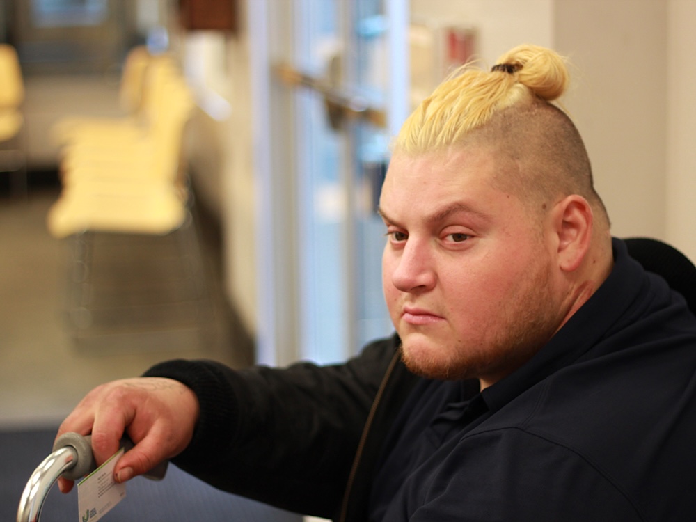 A man with light skin tone and an undercut and bleached hair is seated in a waiting area, with the top of his hand resting on a cane.
