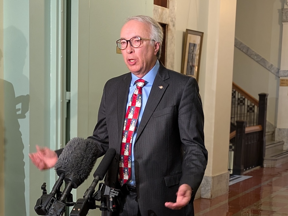 John Rustad, a 60-ish man in a suit with grey hair, stands in front of microphones with his arms spread.