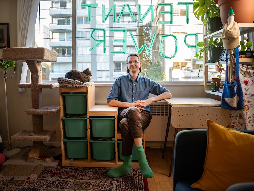 A man with light skin and brown hair sits against a window in a studio apartment. His cat is next to him, and there is a &lsquo;Tenant Power&rsquo; sign written with green tape on the window.