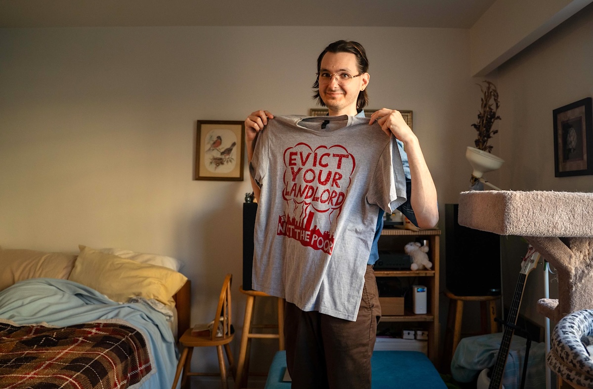 A man in his 30s with light skin, brown hair and glasses holds up a grey T-shirt that says &lsquo;Evict your landlord, not the poor.&rsquo;