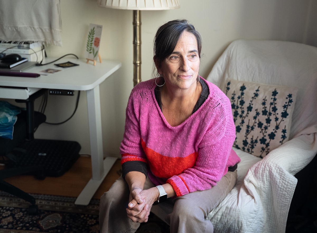 A woman with light skin in her 60s sits in an armchair. She wears a pink and red sweater and has her hair pulled into a ponytail.