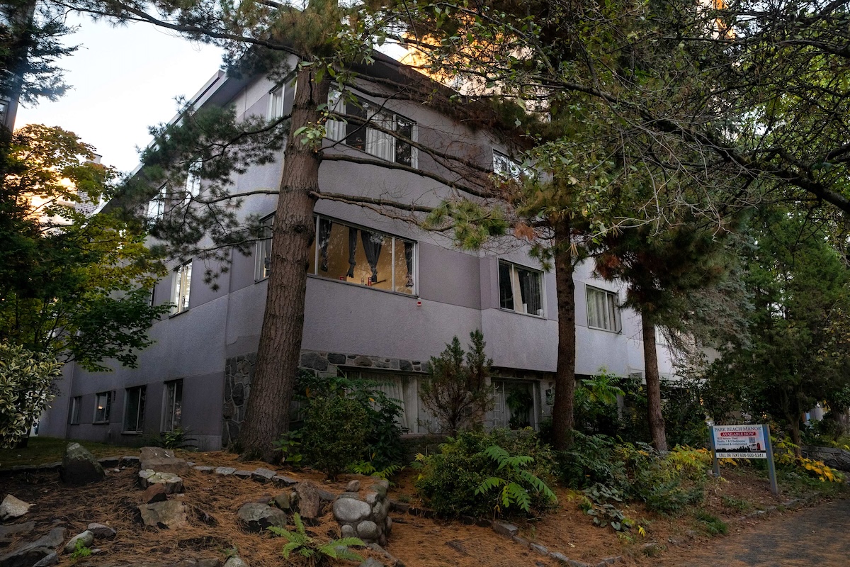 A three-storey apartment building in Vancouver&rsquo;s West End neighbourhood. Several tall evergreen trees stand in front of the building.