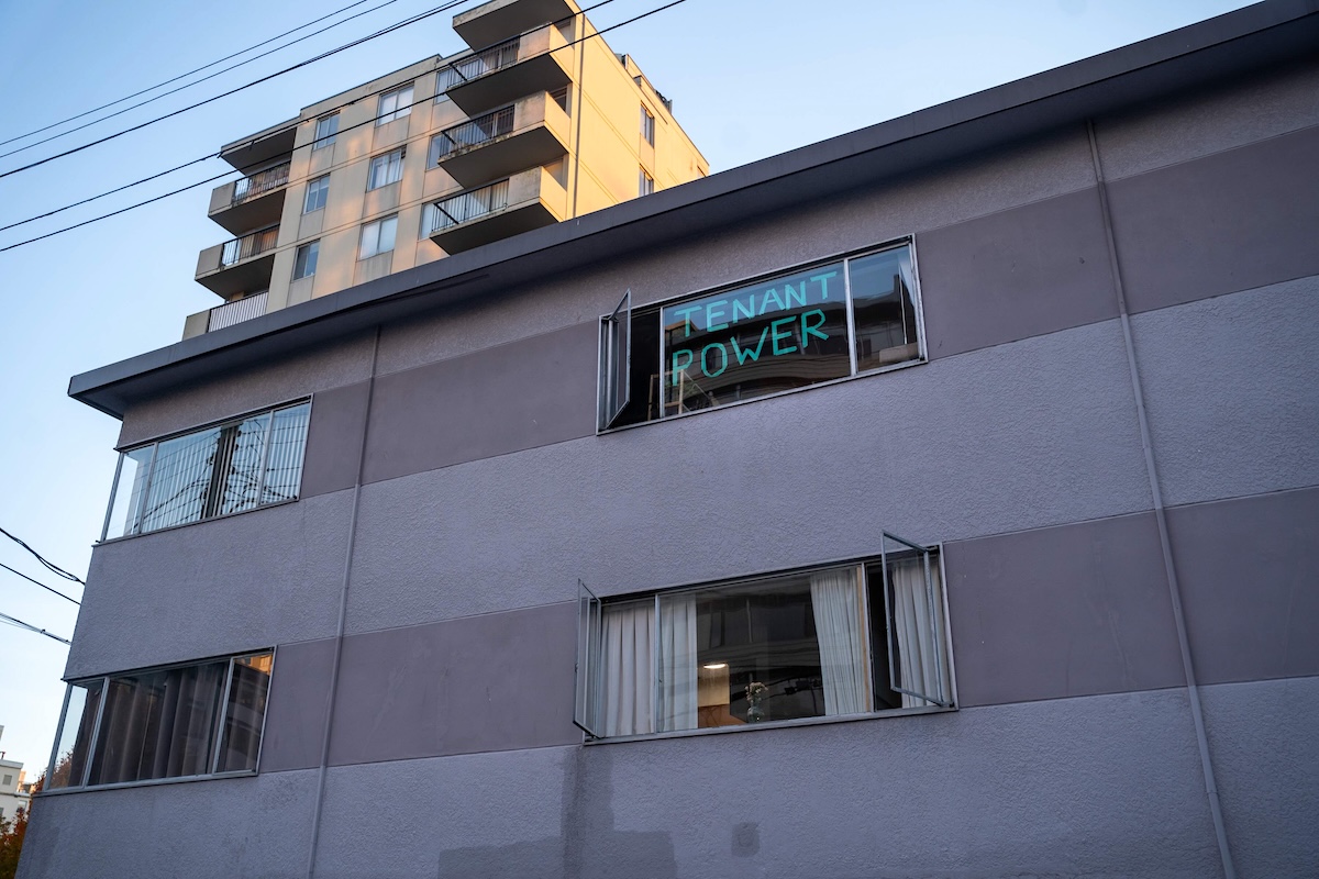 A three-storey apartment building in the West End. On a window overlooking the alley the words &lsquo;Tenant Power&rsquo; can be seen displayed with green tape.