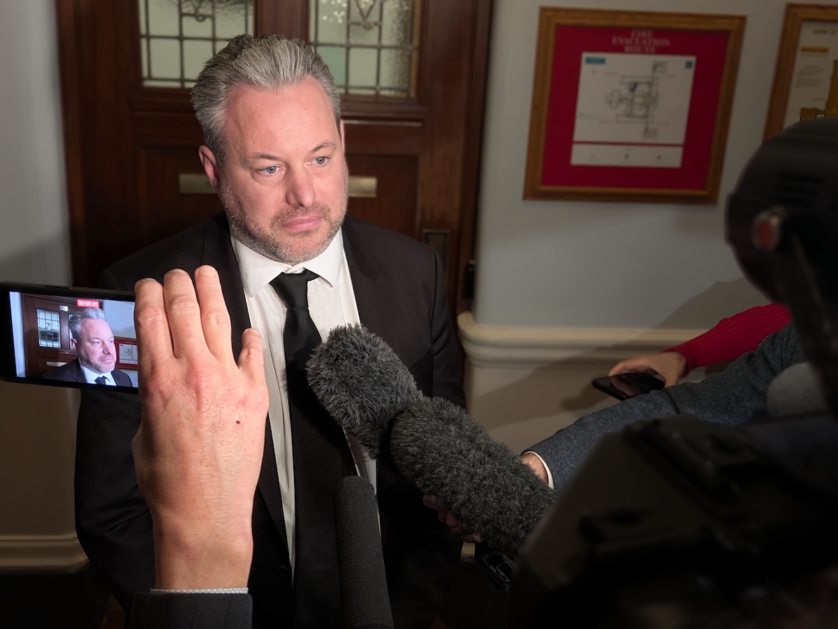 Trevor Halford, a light-skinned man in his mid-40s, looks concerned as reporters ask questions. His image is repeated in a phone screen on the right side of the photo.