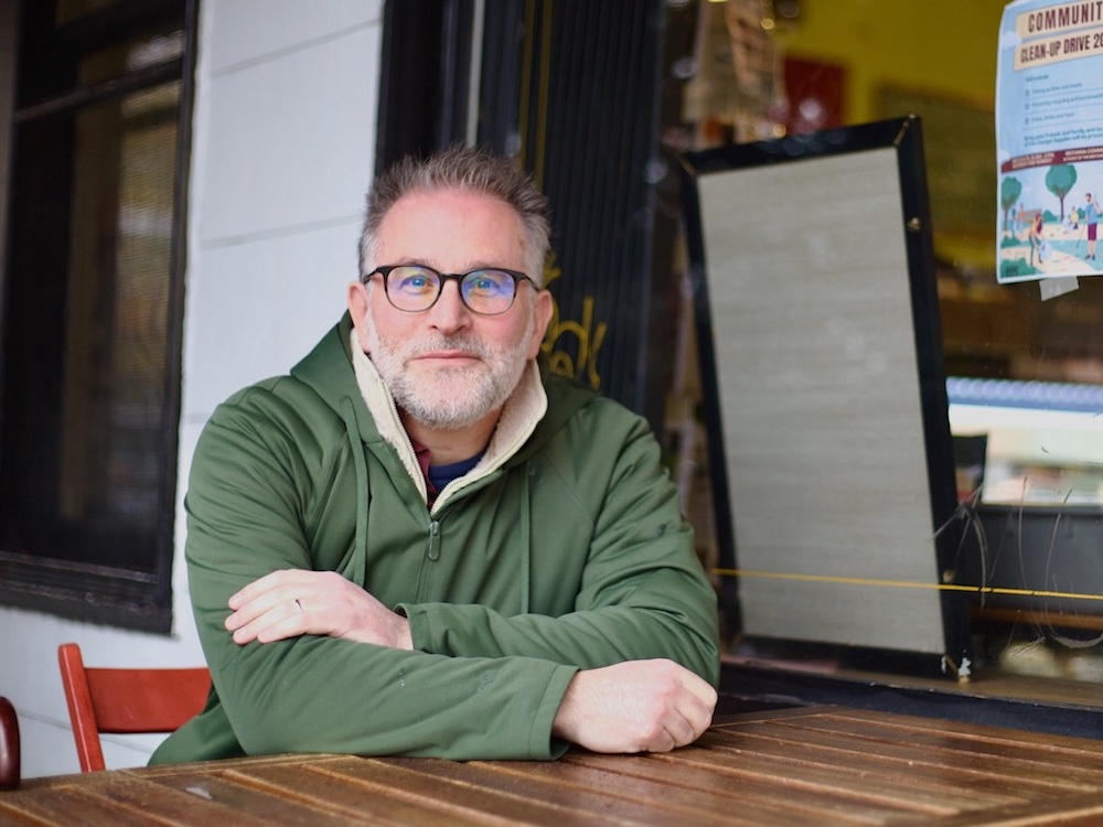A man in his 50s with light skin, glasses and spiky grey hair sits at an outdoor table at a caf&eacute;. He wears a green fleece pullover.