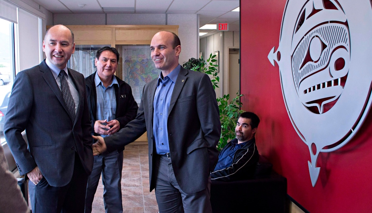 Three men stand and share words while another, seated man watches. A large art piece adorns a nearby wall.