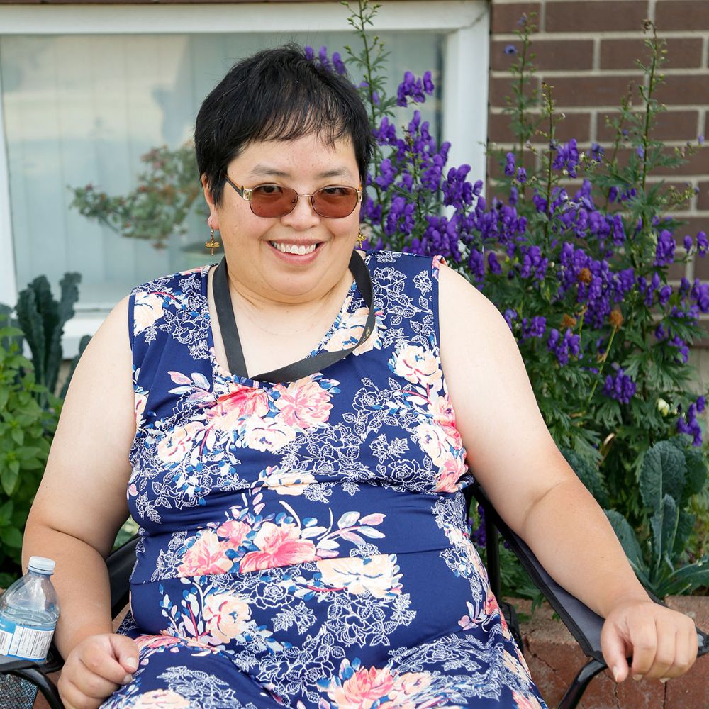 A light-skinned woman with short black hair smiles at the camera in a garden. She is wearing a blue print dress and orange sunglasses.