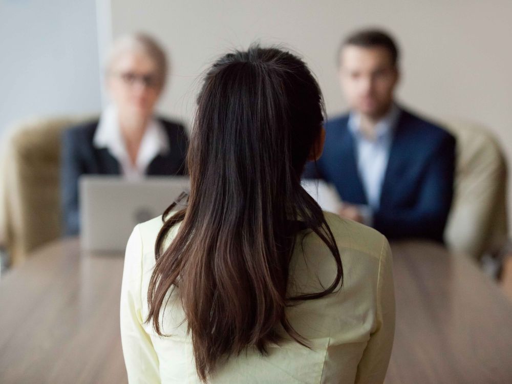 A woman with dark hair in a ponytail sits with her back to the camera, sitting at a desk with two other people facing her.