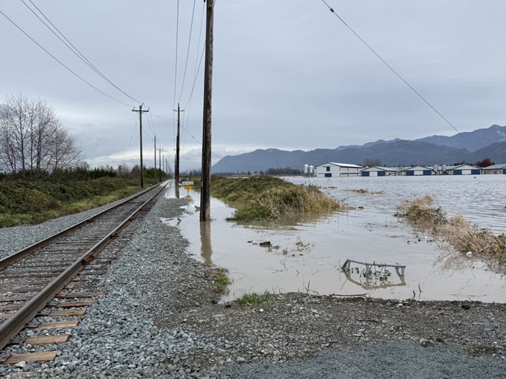 Water creeps towards a rail line across a field in the foreground. In the background, water has already inundated the line.