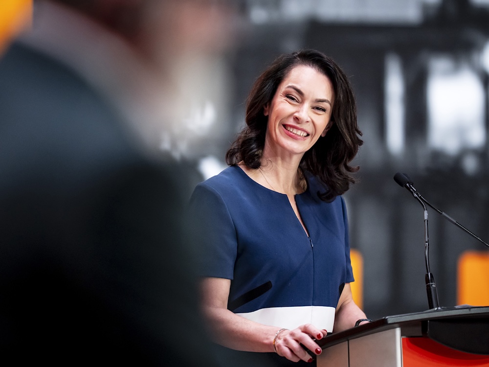 A woman with light skin and shoulder-length brown hair smiles brightly as she stands at a lectern.